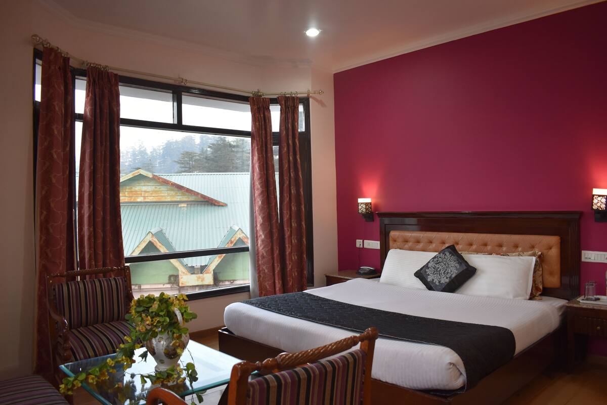 A hotel room with a large bed, a chair, a table with a vase of flowers, and a view of a building outside.