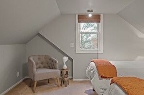 Une chambre avec un plafond en pente, un lit simple agrémenté d’un plaid orange, un fauteuil à capitons, une petite table d’appoint en bois et une fenêtre donnant sur la forêt.