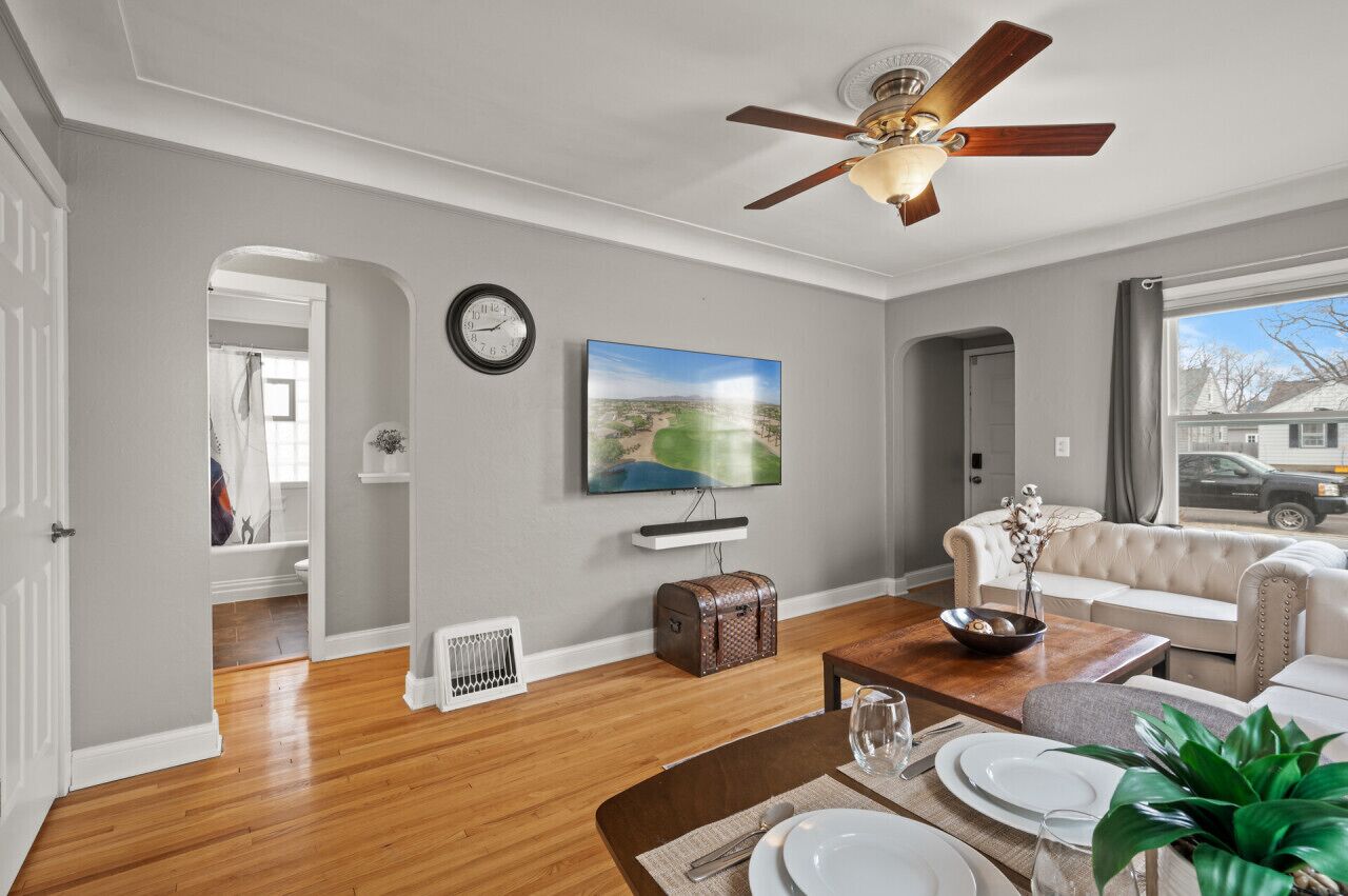 A living room with a ceiling fan, a television mounted on the wall, a sofa, a dining table set for four, and a view of the outside through a window.