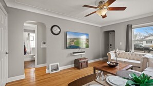 Una sala de estar con ventilador de techo, televisor montado en la pared, sofá, mesa de comedor para cuatro personas y vista al exterior a través de una ventana.