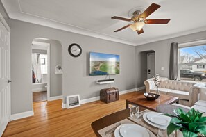 Una sala de estar con ventilador de techo, televisor montado en la pared, sofá, mesa de comedor para cuatro personas y vista al exterior a través de una ventana.