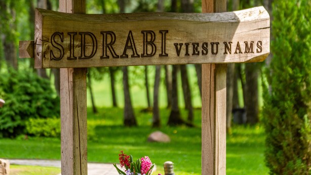 Un panneau en bois portant l’inscription « SIDRABI VIESUNAMS » dans une zone boisée.