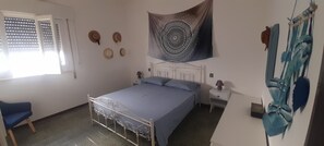 A bedroom with a bed, a window with blinds, a bedside table with a lamp, and a tapestry on the wall.