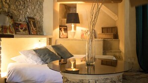 A cozy bedroom with a stone wall, a bed with white and gray bedding, a bedside table with a lamp, and a vase with branches. - In Provence Hôtel (Venelles)