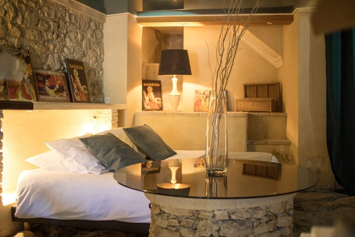 A cozy bedroom with a stone wall, a bed with white and gray bedding, a bedside table with a lamp, and a vase with branches.