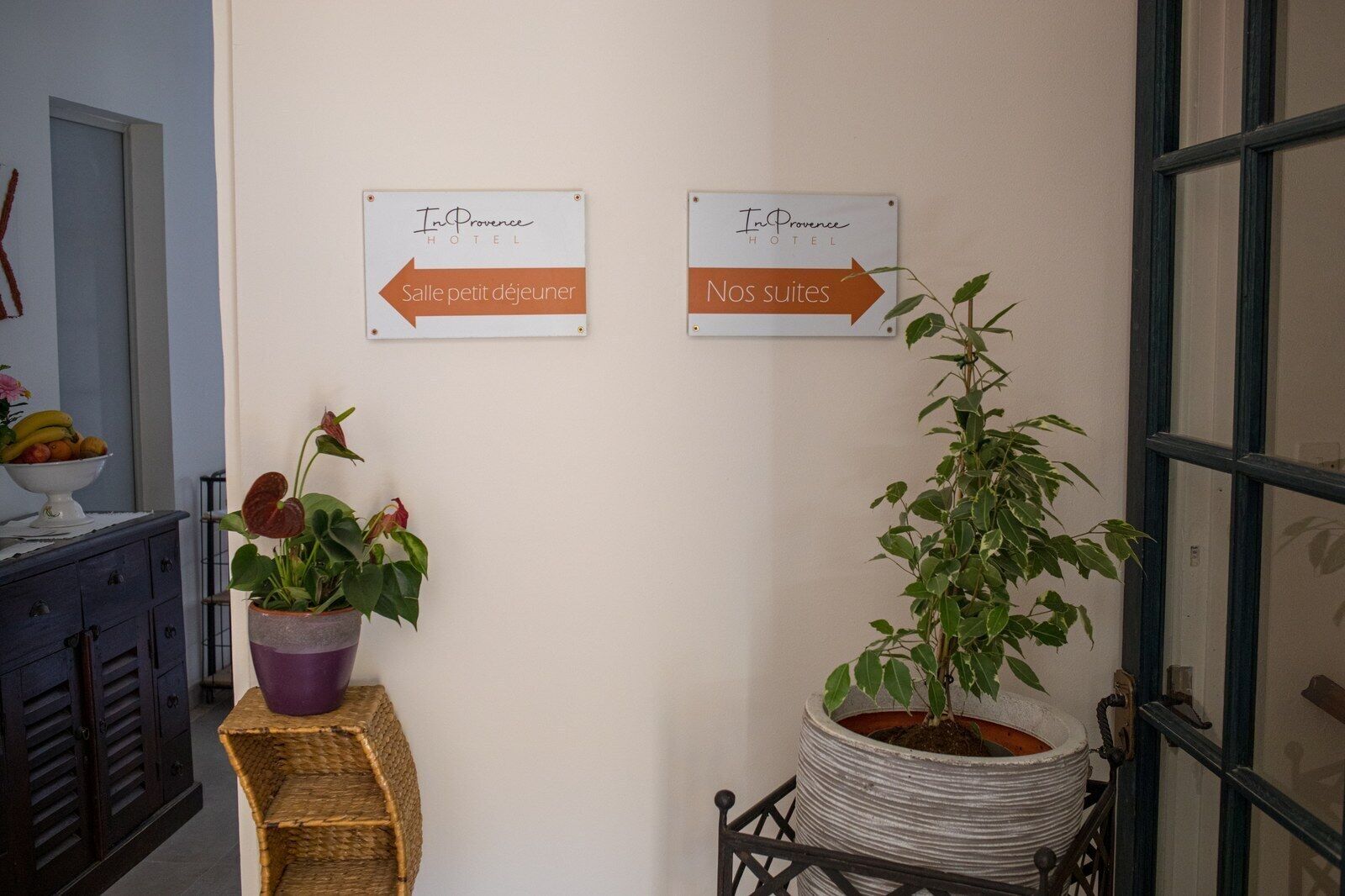 A hallway with two signs indicating directions to the hotel lobby and rooms. There are potted plants on a stand and a wicker basket.