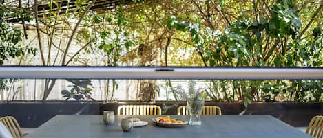 An outdoor dining area with a table set for two, surrounded by chairs, and a view of greenery and a building in the background.
