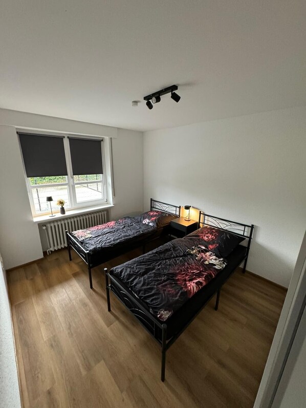 A small, well-lit room with two single beds, a window with blinds, a radiator, and a ceiling light fixture.