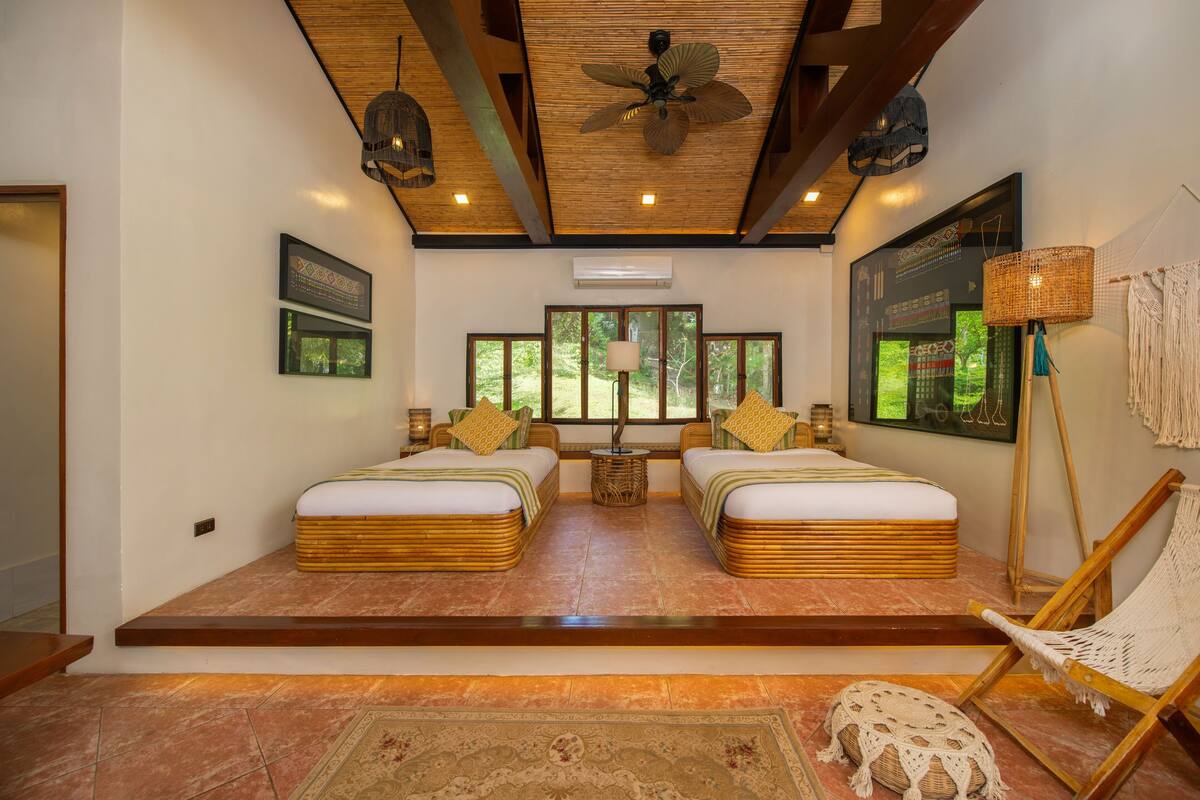 A room with two beds, a ceiling fan, a large window, a woven chair, and a rug.