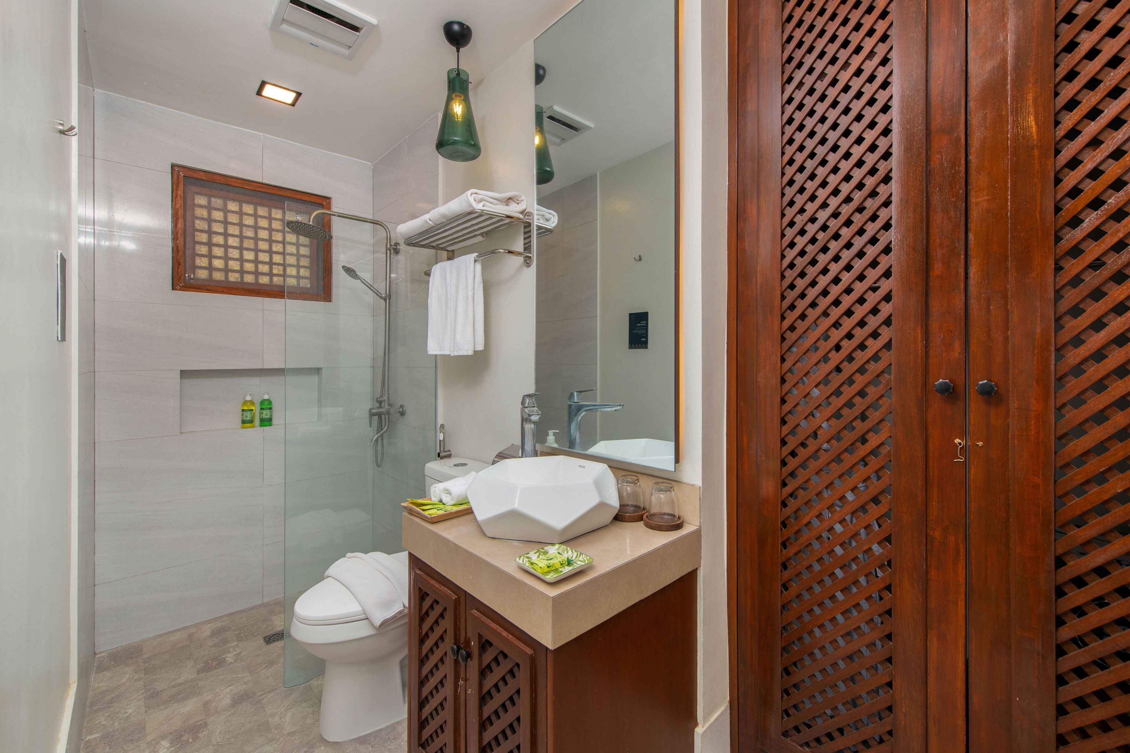 A modern bathroom with a glass shower enclosure, a white toilet, a beige vanity with a large mirror, and a wooden door with a lattice pattern.