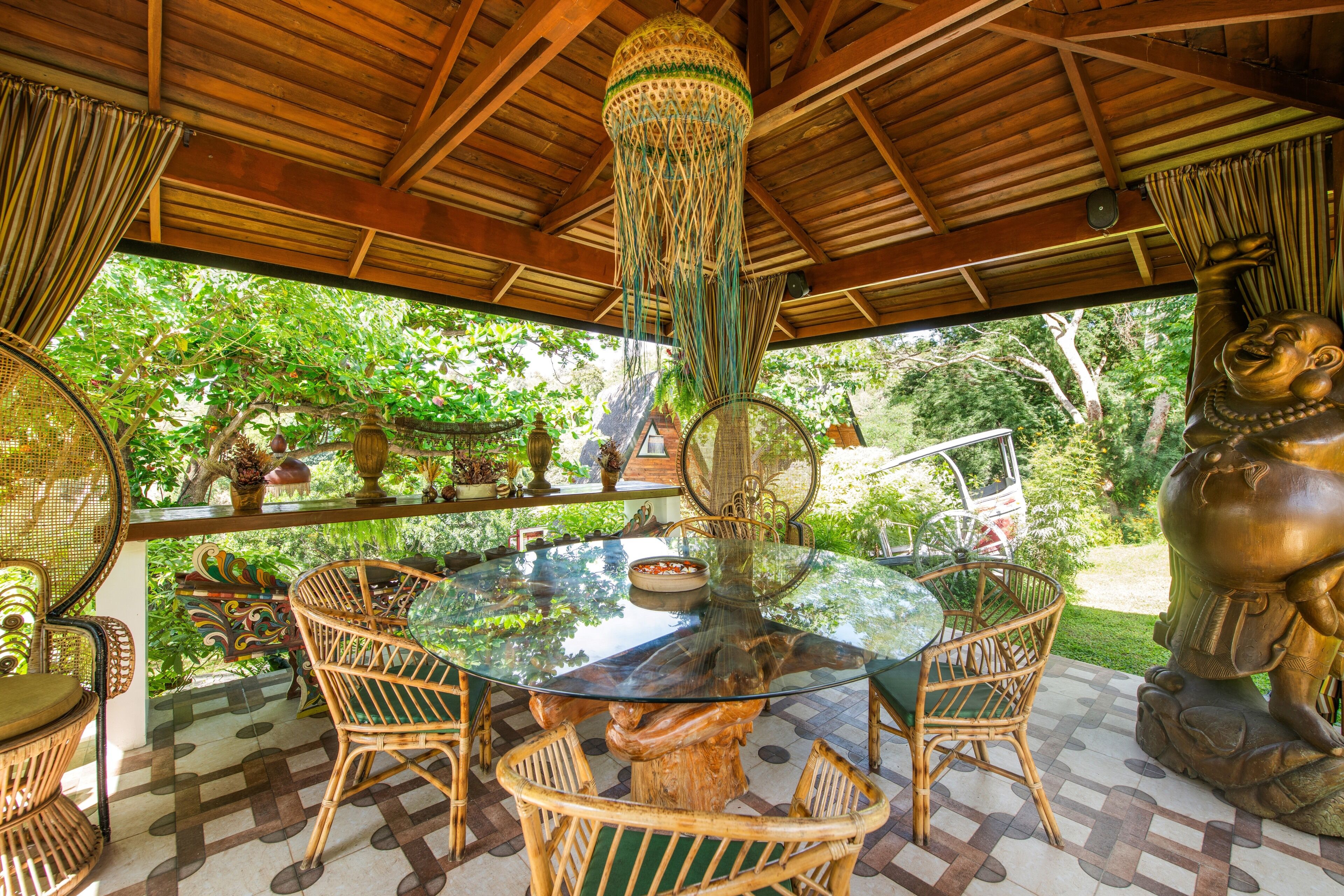 A covered outdoor dining area with a glass table surrounded by wicker chairs, a hanging light fixture, and a statue.