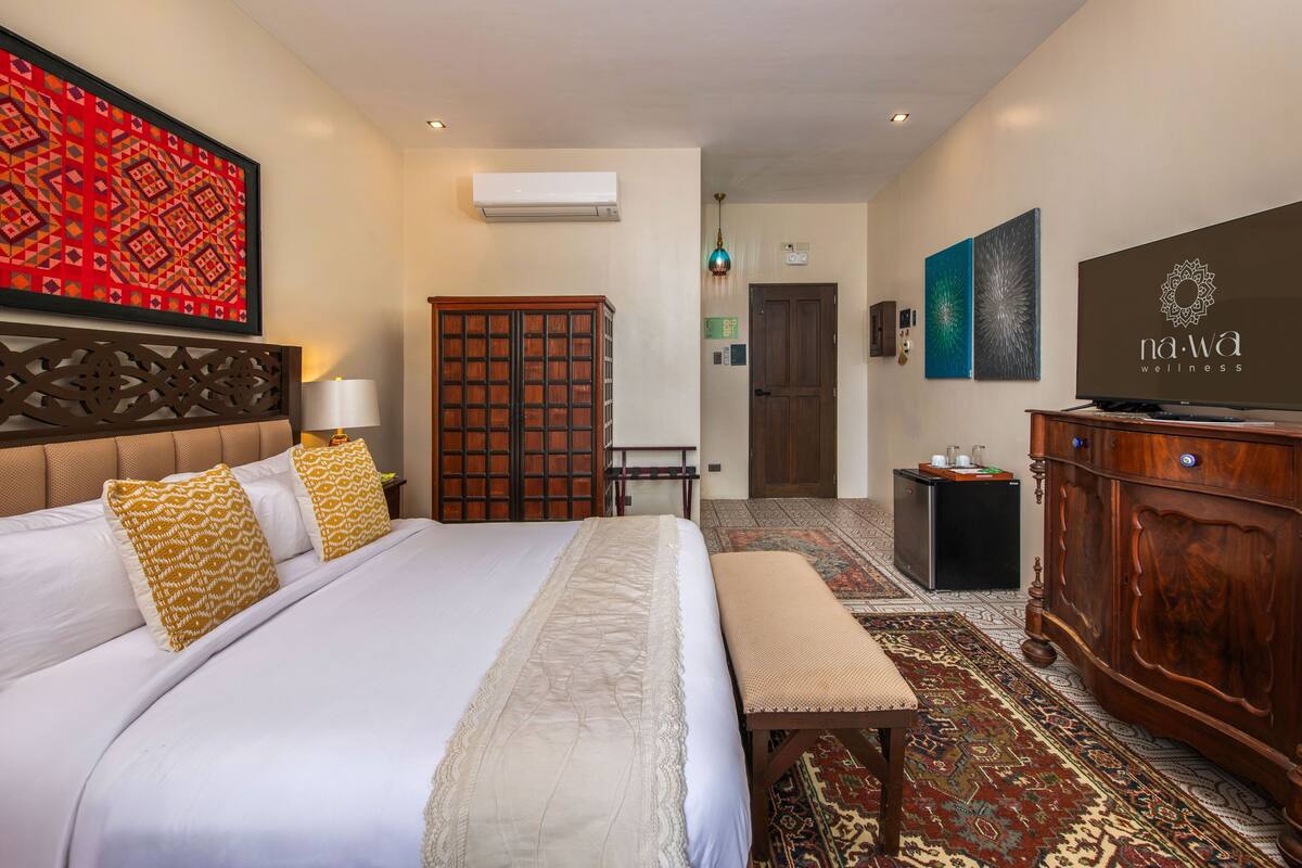 A hotel room with a large bed, a wooden wardrobe, a television, and a patterned rug.