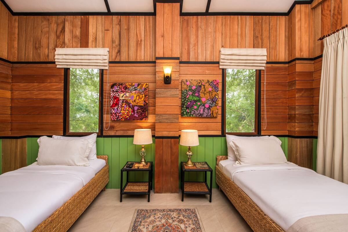 A room with two beds, wooden walls, artwork, lamps, and a view of trees.
