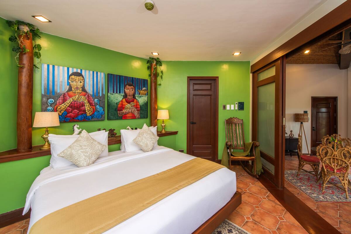 A hotel room with a large bed, green walls, two paintings, a wooden door, and a chair.