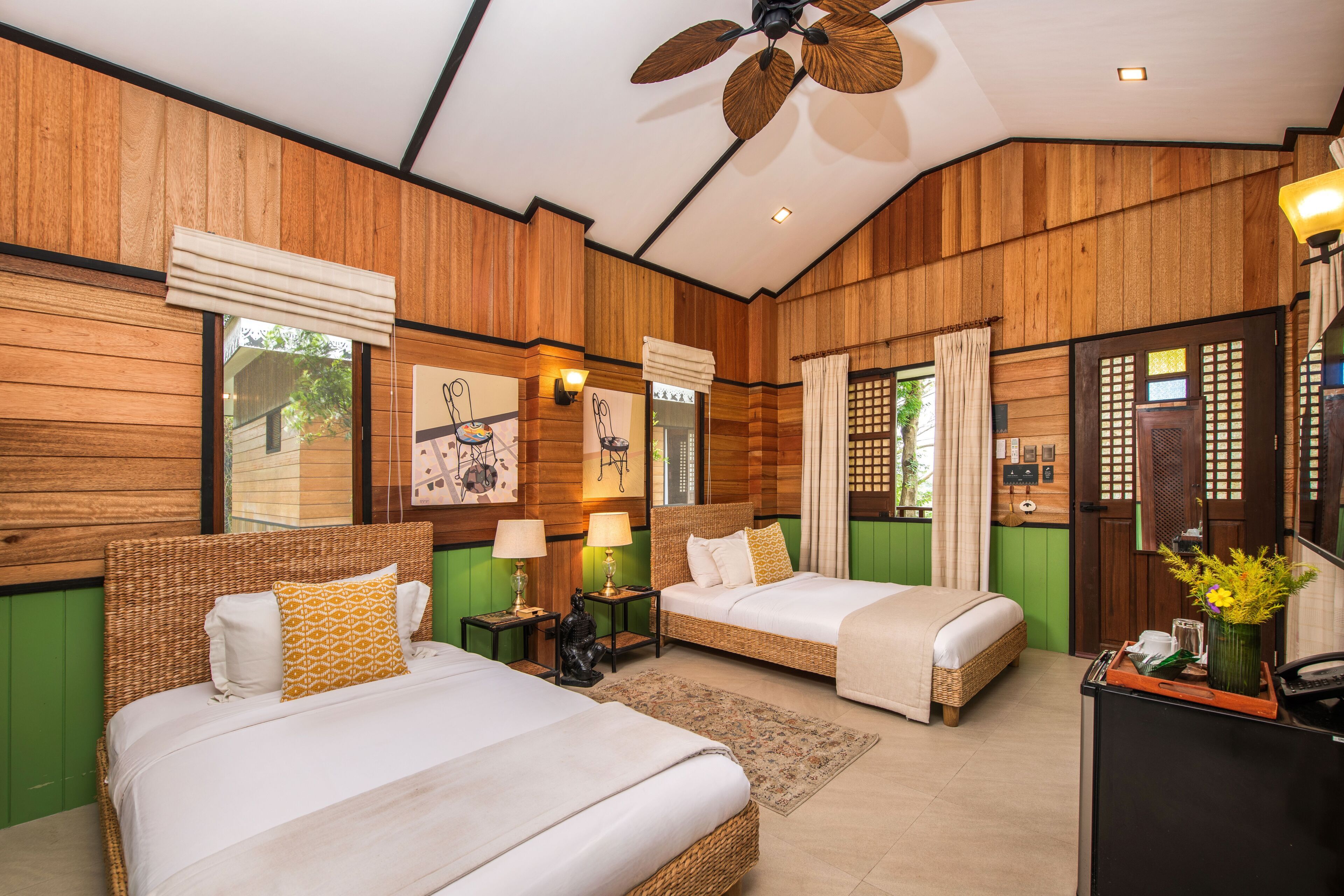 Habitación de hotel con dos camas, paneles de madera, ventilador de techo y un escritorio con un florero.
