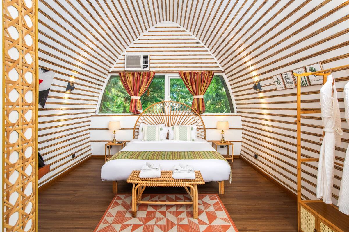 A room with a bed, a window with curtains, a bedside table, a wall-mounted air conditioner, and a wooden floor.