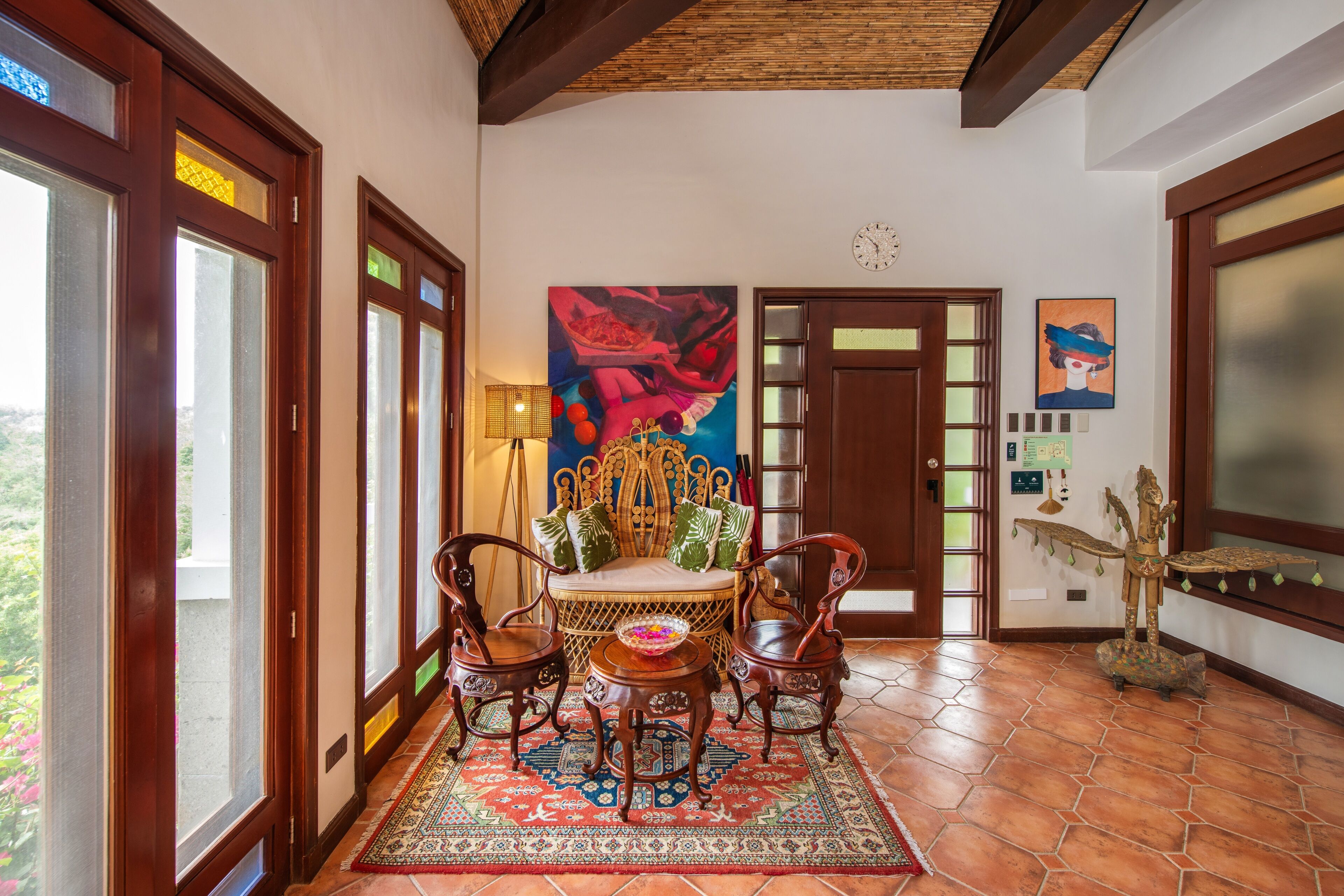 A room with a wicker sofa, two wooden chairs, a patterned rug, a decorative statue, and a large painting on the wall.