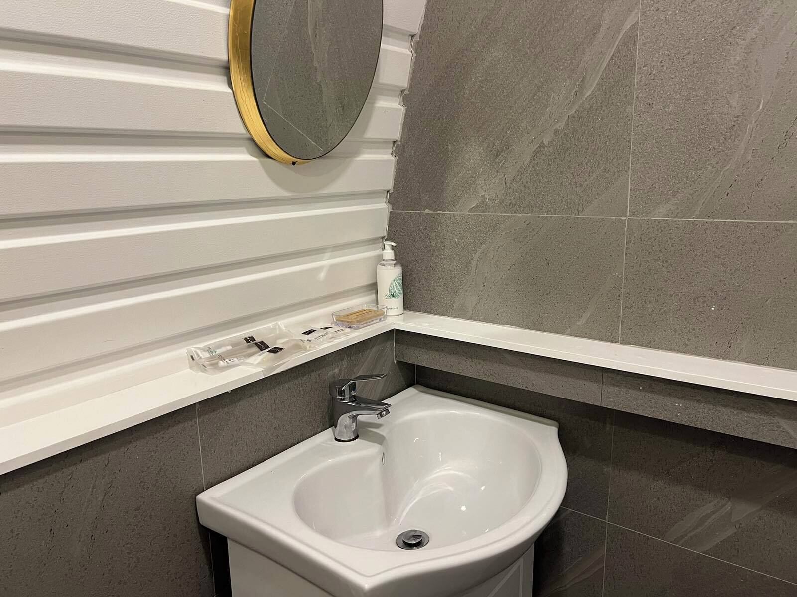 Een badkamer met een wit wastafeltje op een zuil, een spiegel met gouden lijst en grijze betegelde wanden.