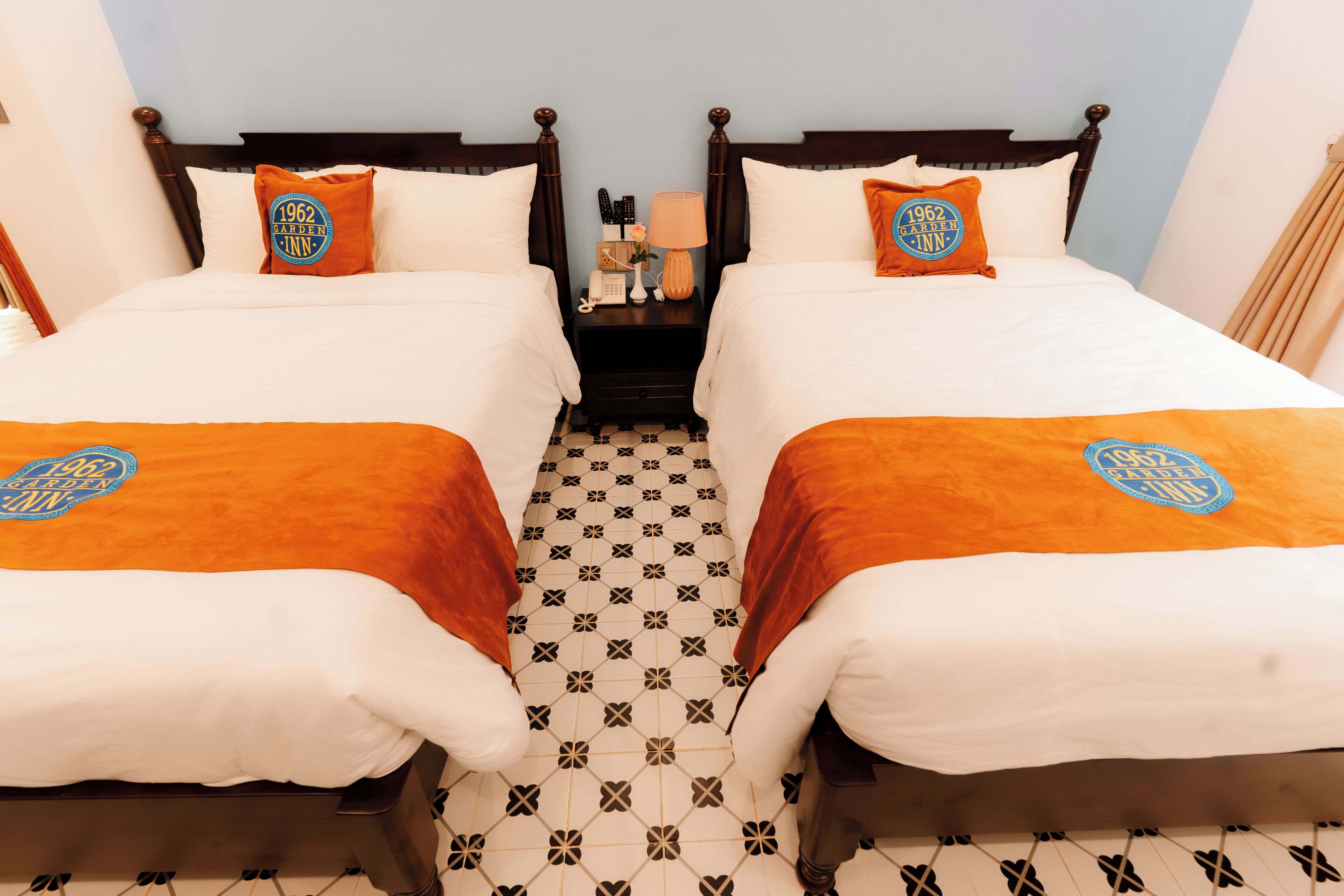 Quarto de hotel com duas camas, cada uma com cabeceira e travesseiro combinando, uma mesa de cabeceira com abajur e piso estampado.
