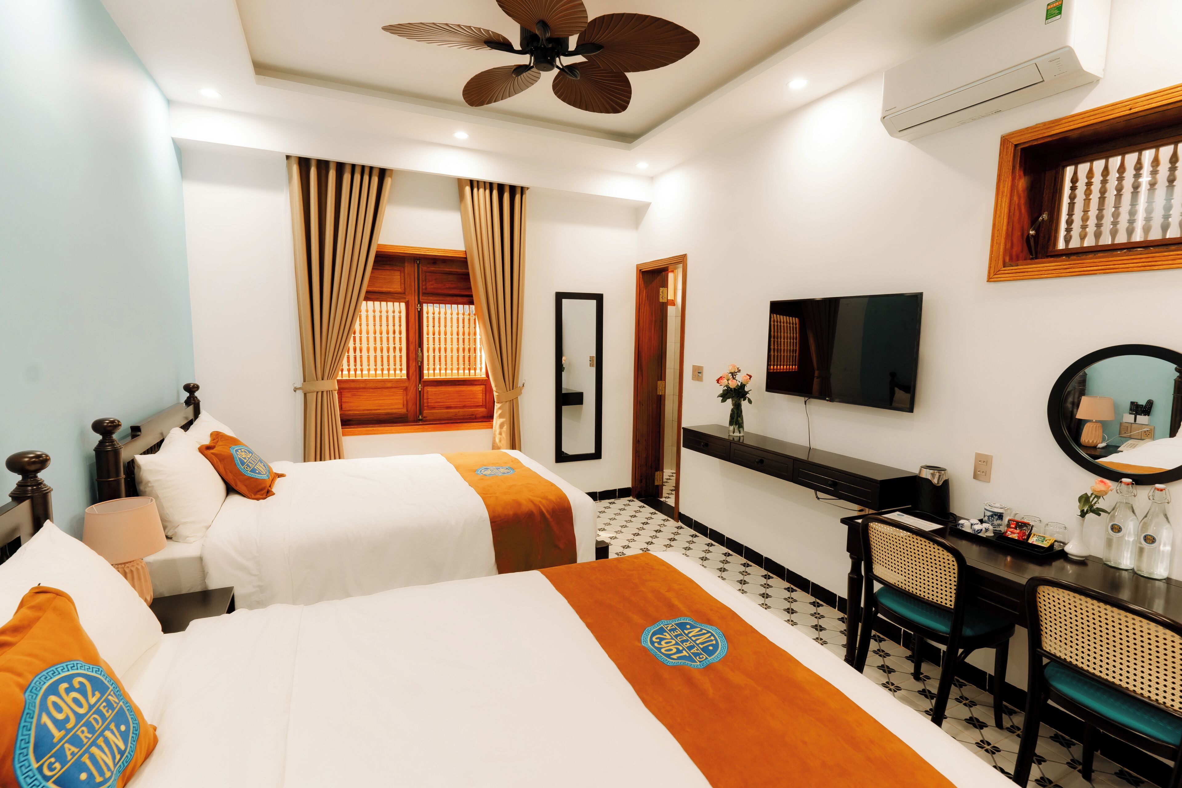 A hotel room with two beds, a desk with chairs, a television, and a ceiling fan.