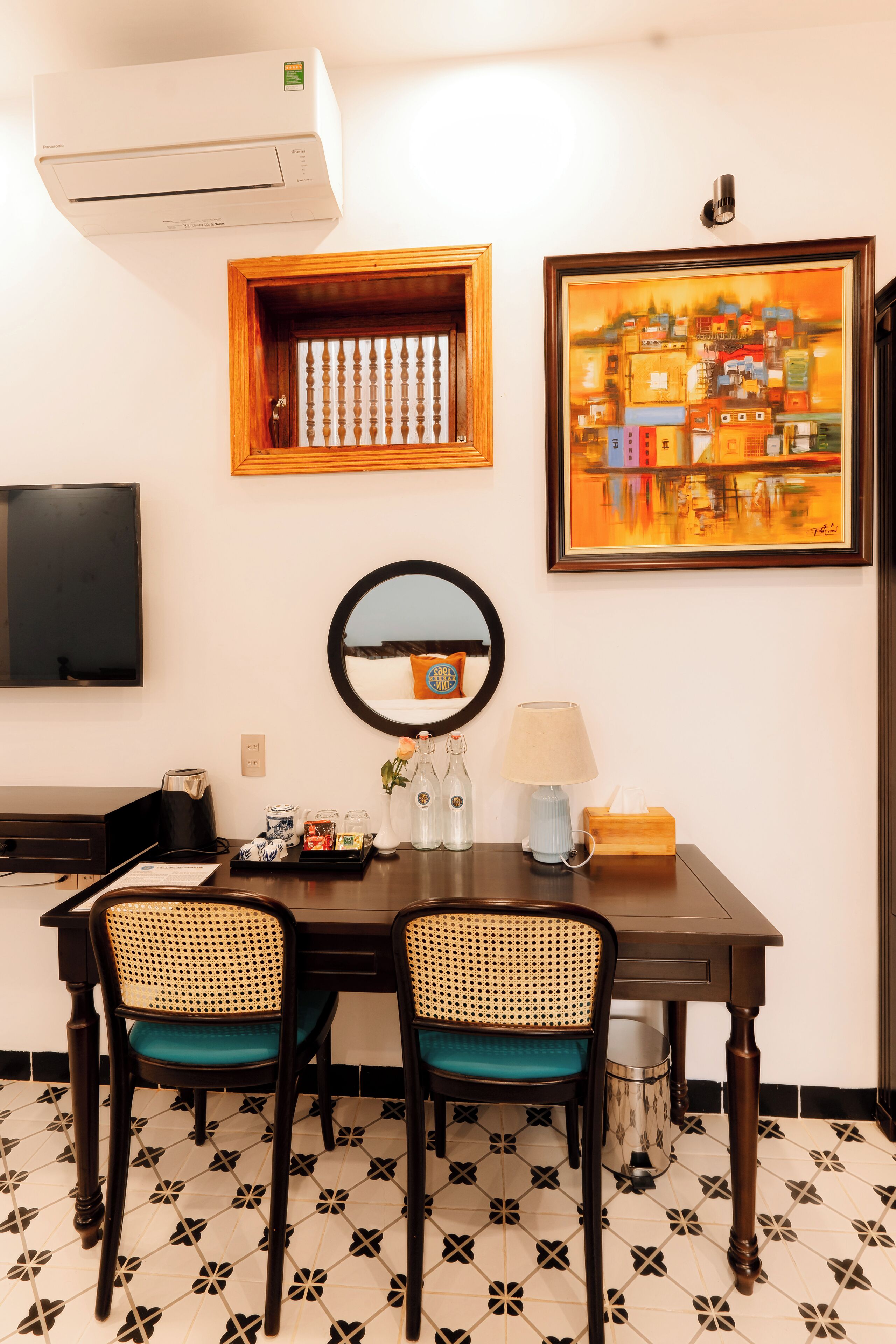 A hotel room with a dining table, two chairs, a mirror, a painting, a television, and an air conditioner.
