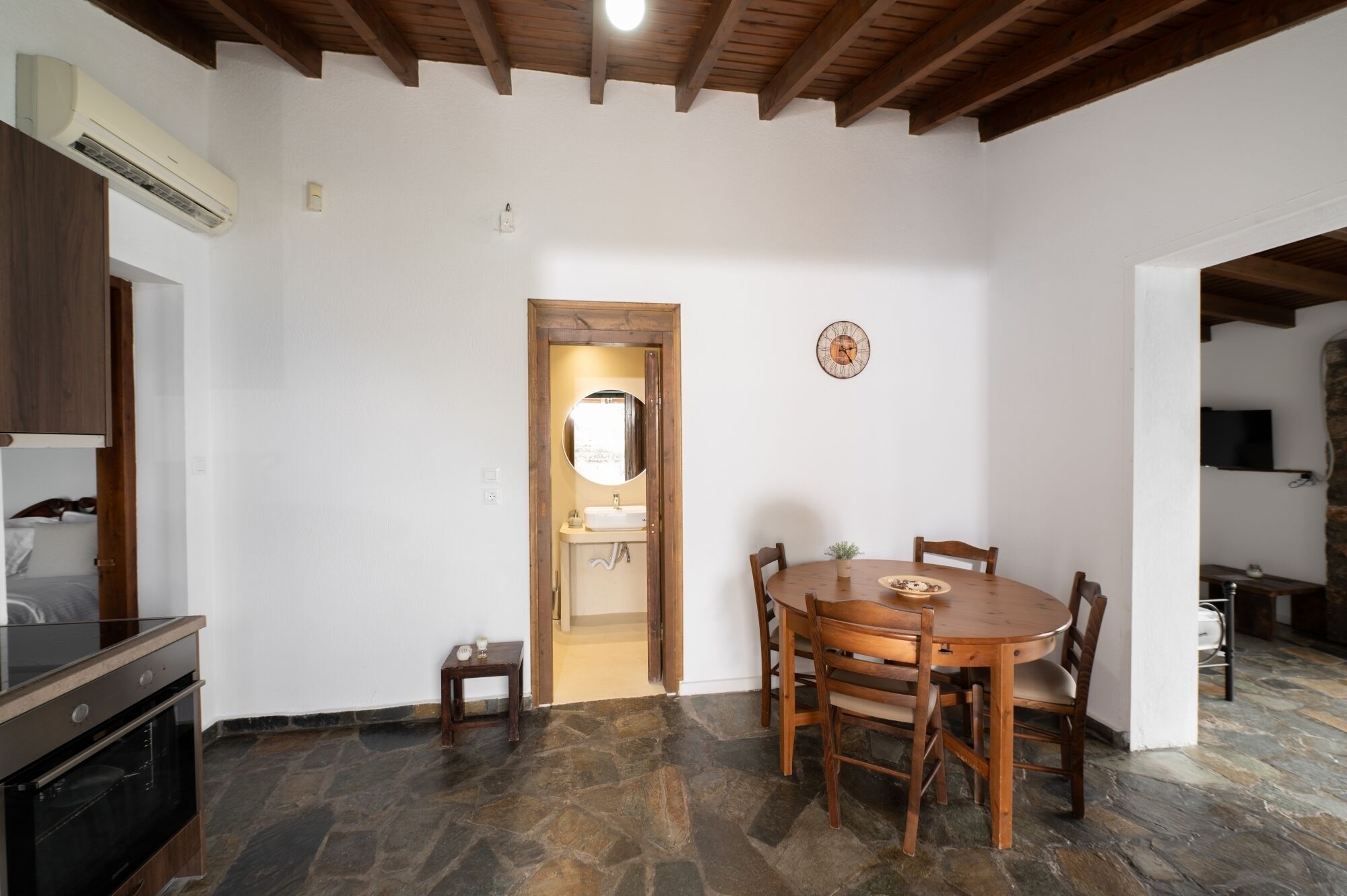 Uma área de jantar com uma mesa de madeira redonda e cadeiras, uma pequena mesa lateral, um relógio na parede e um forno embutido.