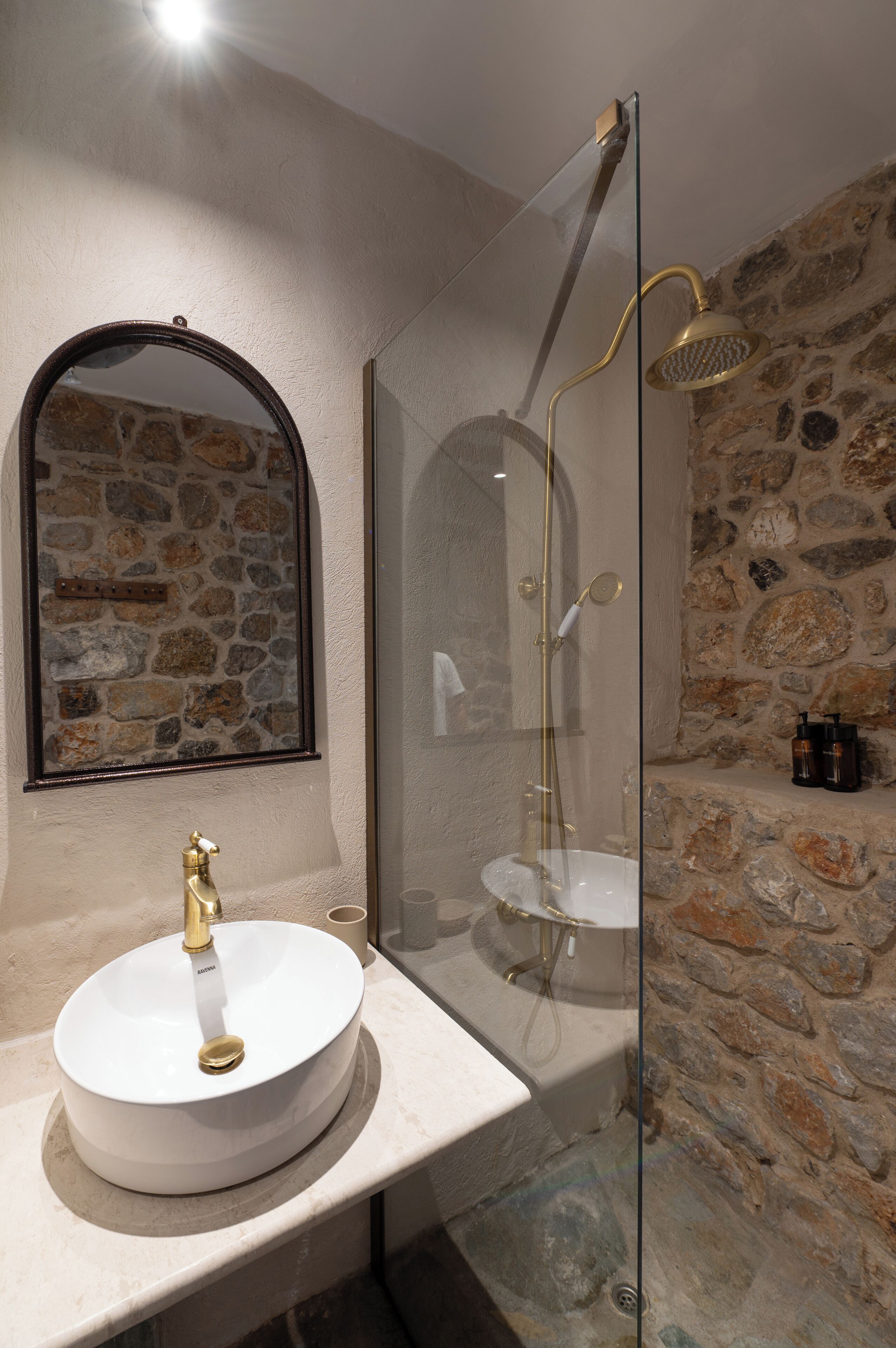 A bathroom with a stone wall, a round sink with a gold faucet, and a glass-enclosed shower area.