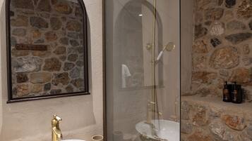 A bathroom with a stone wall, a round sink with a gold faucet, and a glass-enclosed shower area.
