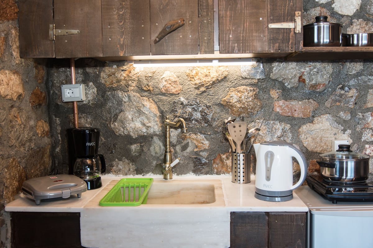 A kitchen with a stone wall, a sink, a coffee maker, a kettle, and a food warmer.