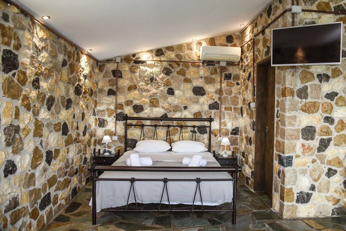 A stone-walled bedroom with a metal bed frame, two bedside tables with lamps, and a flat-screen TV mounted on the wall.