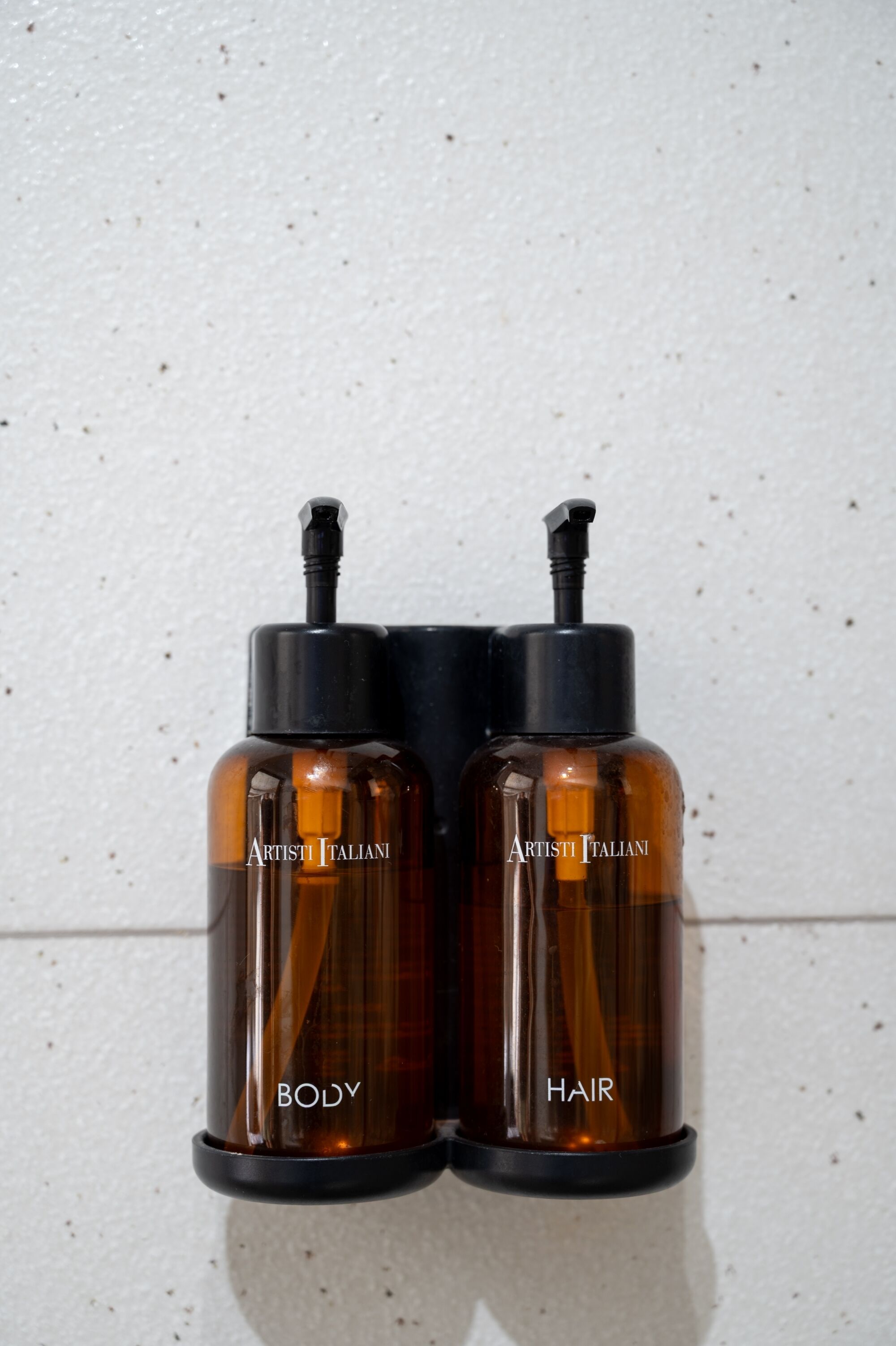 Dos botellas marrones con las etiquetas 'ARTISTI ITALIANI BODY' y 'ARTISTI ITALIANI HAIR' sobre un muro blanco.