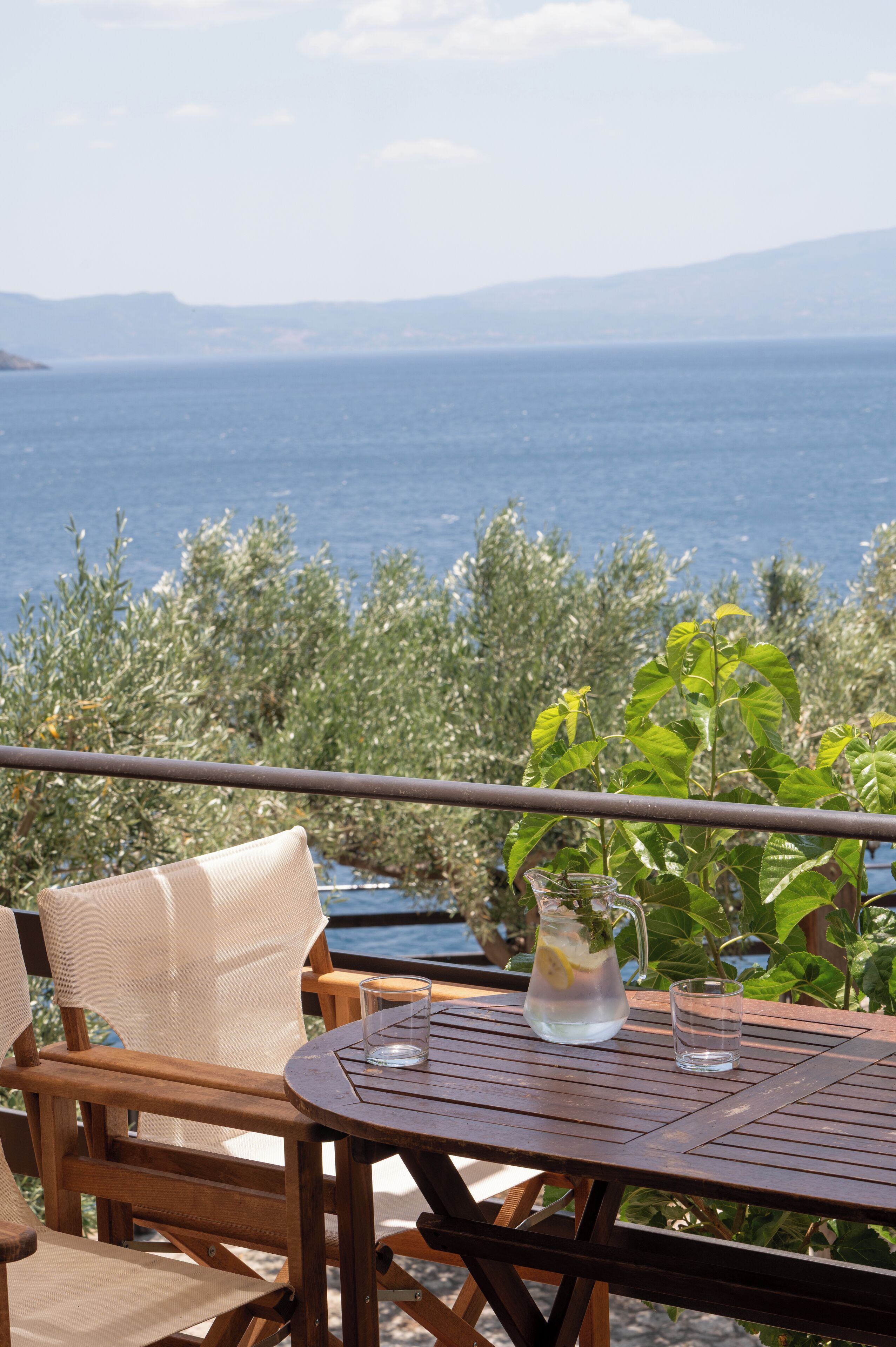 A wooden table with a pitcher and glasses on a balcony overlooking a body of water and greenery.