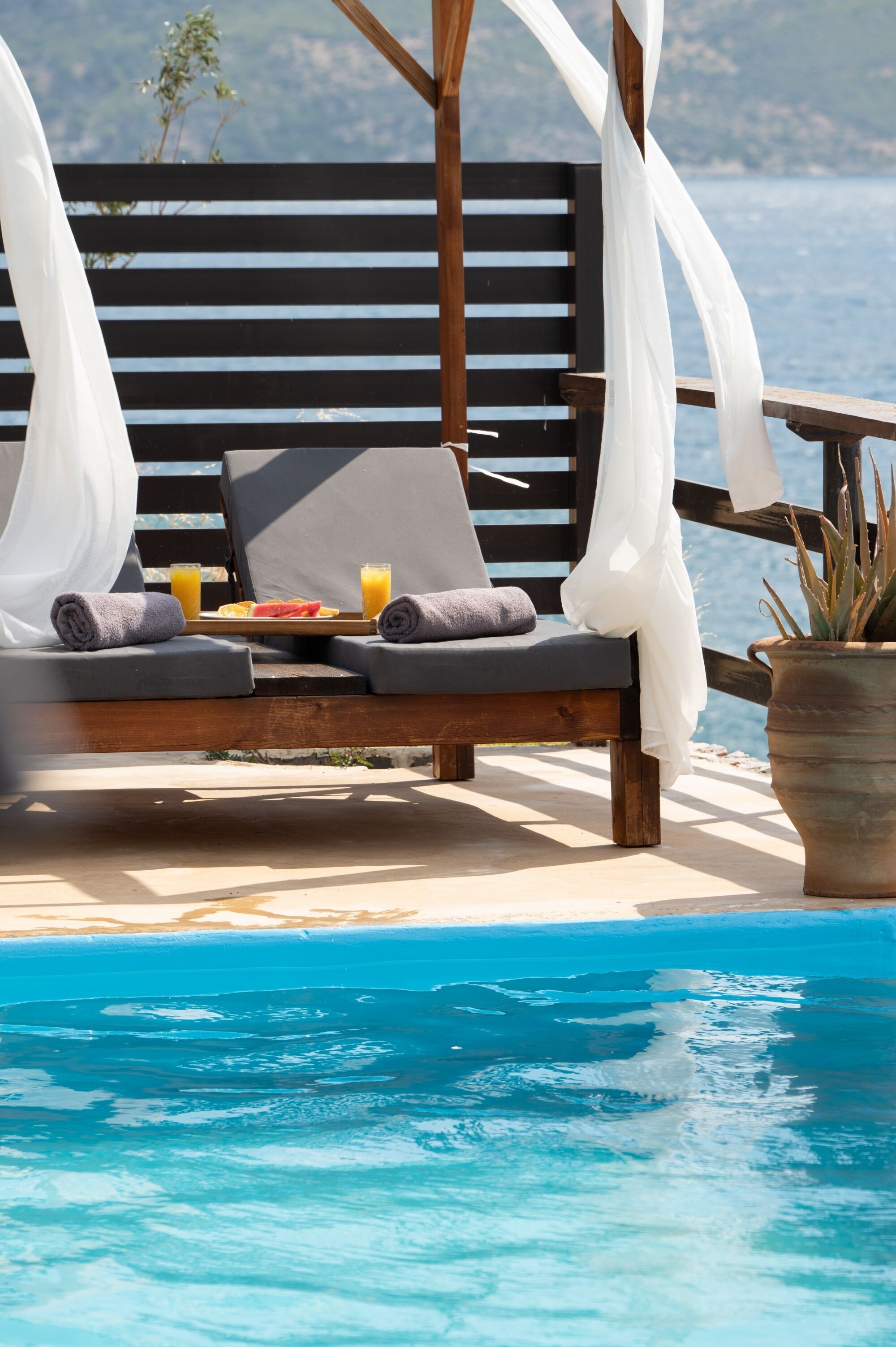 Un fauteuil de jardin en bois avec un coussin, un plateau avec des fruits et du jus, et une plante en pot près d’une piscine.