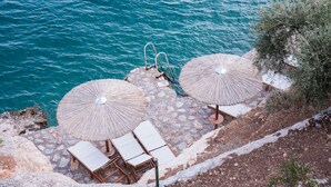 Een terras aan de kust met stenen vloer, twee parasols en loungestoelen met uitzicht op zee.