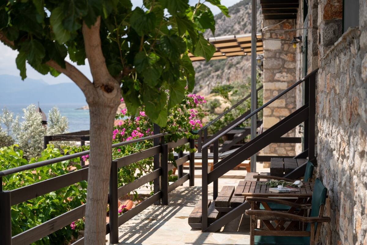 A wooden staircase leading to a terrace with a view of the sea, surrounded by greenery and a stone wall.