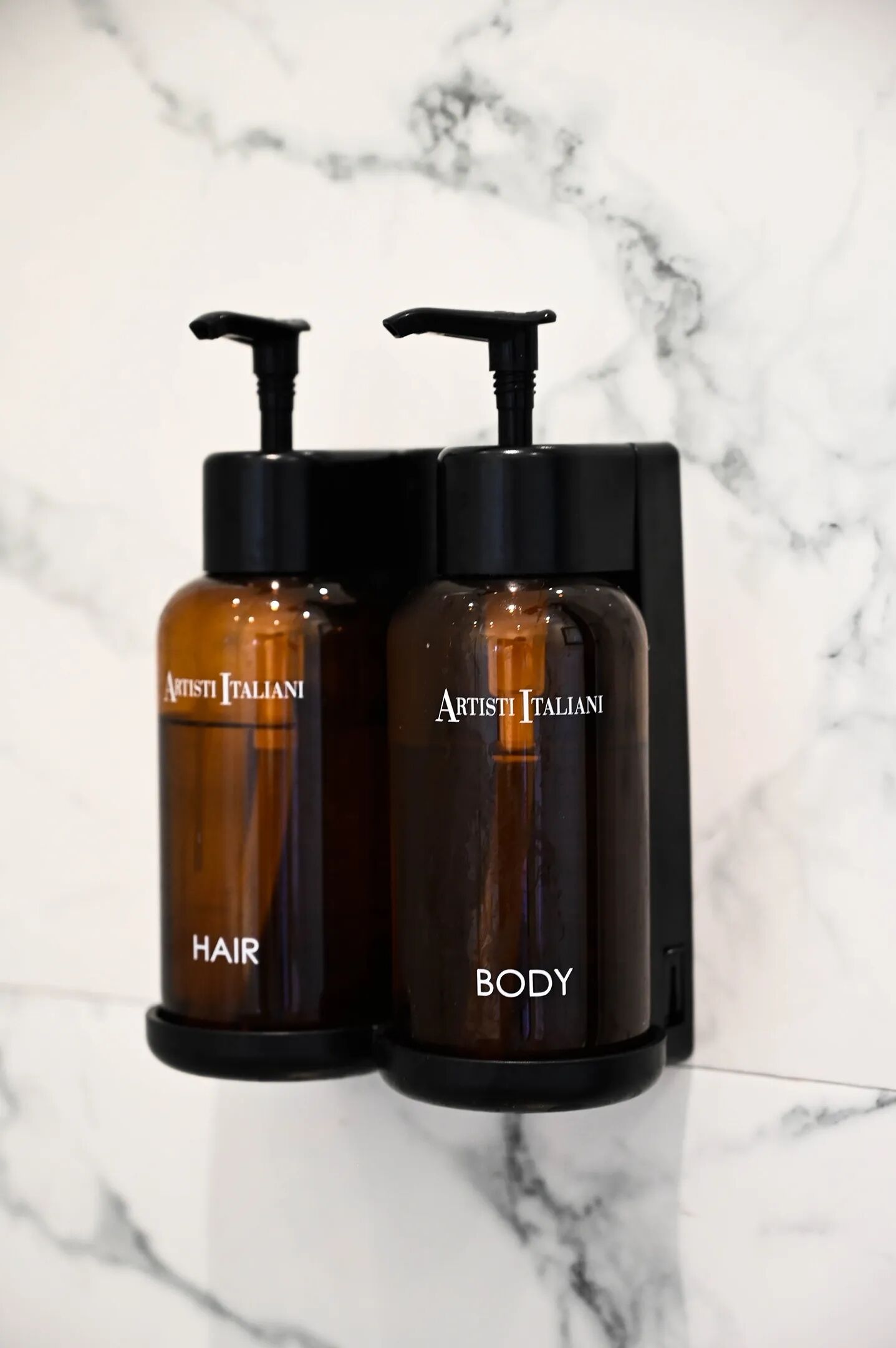 Zwei braune Flaschen mit schwarzen Pumpen, beschriftet mit 'ARTISTI ITALIANI HAIR' und 'ARTISTI ITALIANI BODY', auf einer weißen Marmorplatte angebracht.