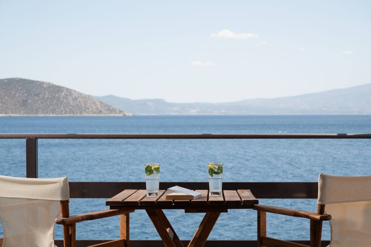 A wooden table with two glasses of water and a book, overlooking a body of water with distant hills.