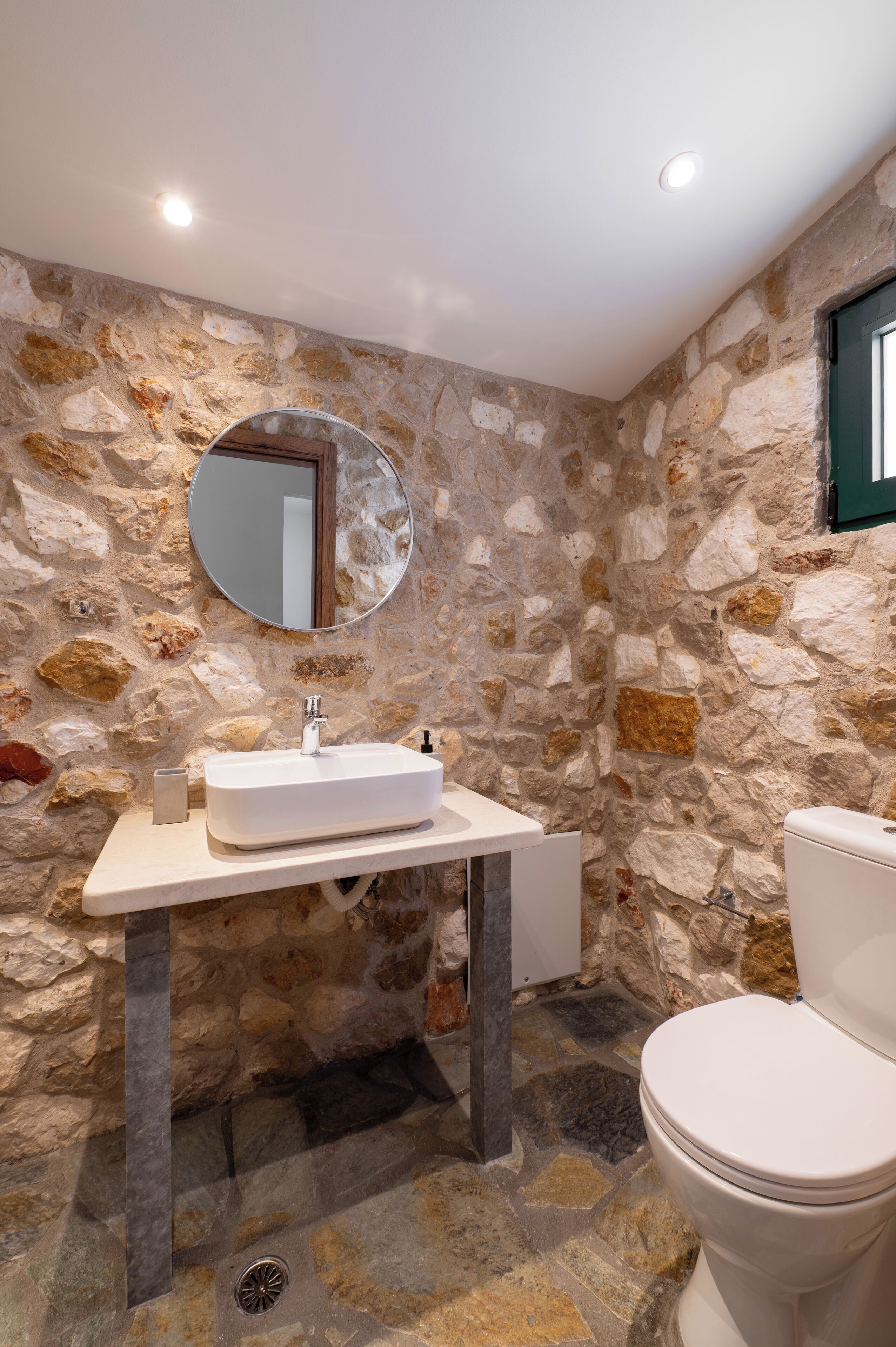 Ein Badezimmer mit Steinwand, rundem Spiegel, weißem Waschbecken und Toilette.