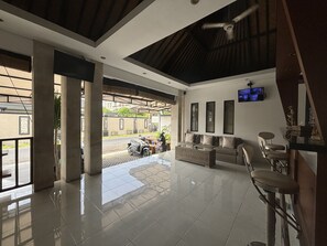 A spacious lobby with a tiled floor, a seating area with a sofa and chairs, a television mounted on the wall, and a motorcycle parked outside.