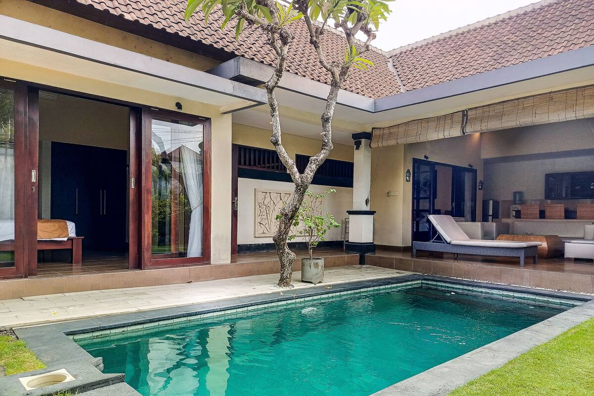 Une piscine avec une terrasse carrelée, un espace salon couvert avec un canapé et une porte donnant sur une pièce où se trouve un lit.