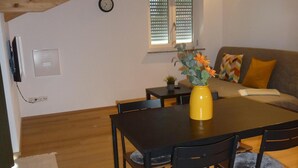 A dining area with a wooden table and chairs, a yellow vase with flowers, and a window with blinds. - Ferienwohnung Seeblick, 1-4 Pers., 1 Sep. Schlafzimmer, Südbalkon (Chieming)