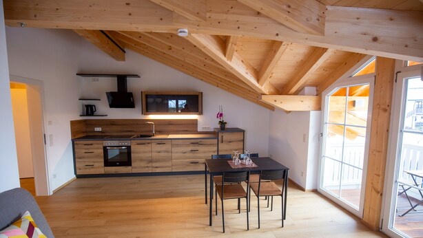 A modern kitchen with wooden beams, a dining area with a table and chairs, and a large window providing natural light. - Ferienwohnung Seeblick, 1-4 Pers., 1 Sep. Schlafzimmer, Südbalkon (Chieming)