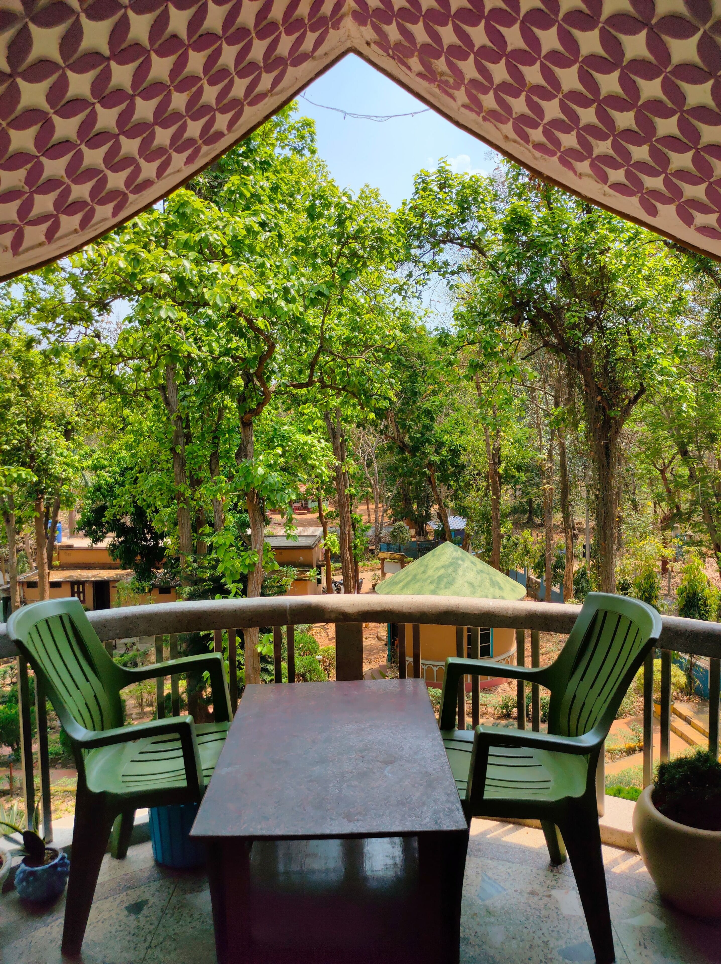 A covered outdoor seating area with a table and chairs, surrounded by trees and a view of buildings in the background.