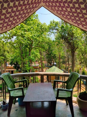 A covered outdoor seating area with a table and chairs, surrounded by trees and a view of buildings in the background. - Rimil EcoTourism (Khatra)