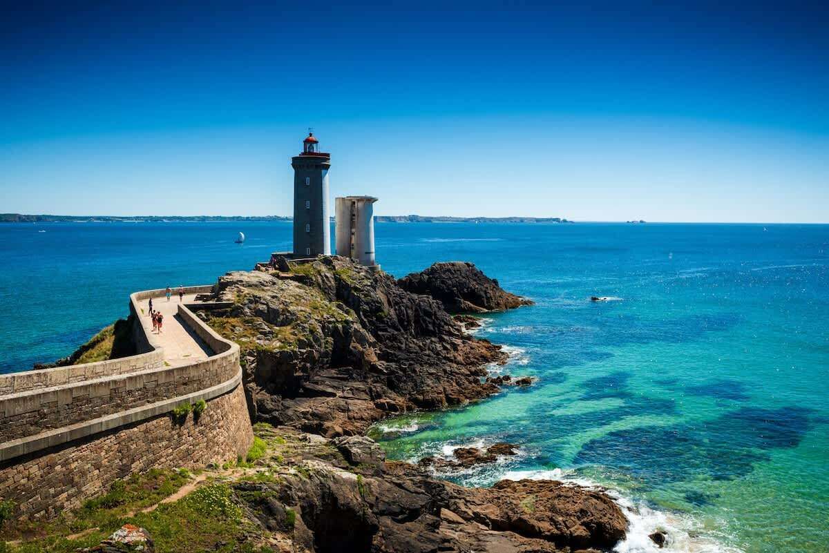 Un faro costero con un camino de piedra que lleva a él, con vista al mar azul y claro.