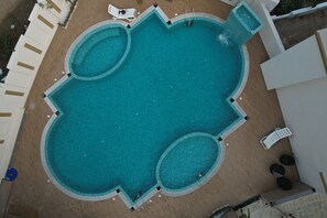 A large, clear swimming pool with a unique shape, surrounded by a white deck and lounge chairs.