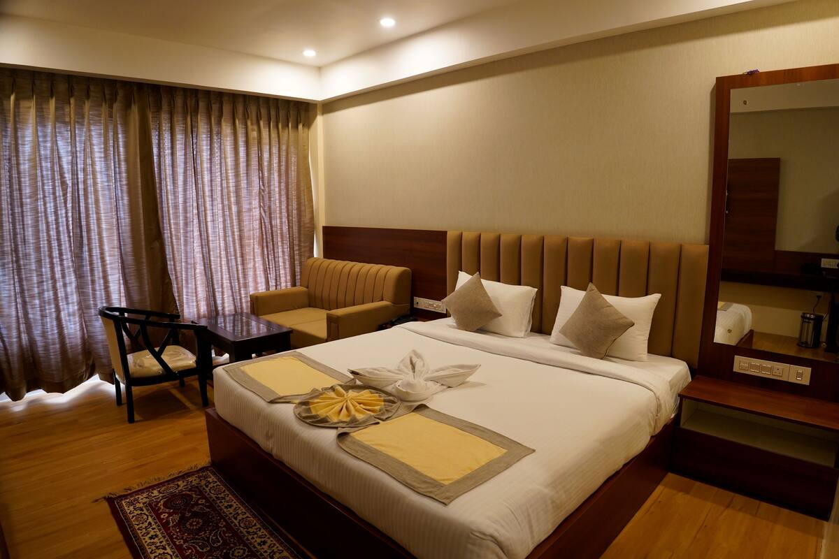 A hotel room with a large bed, a wooden headboard, a bedside table, a chair, a mirror, and a window with curtains.