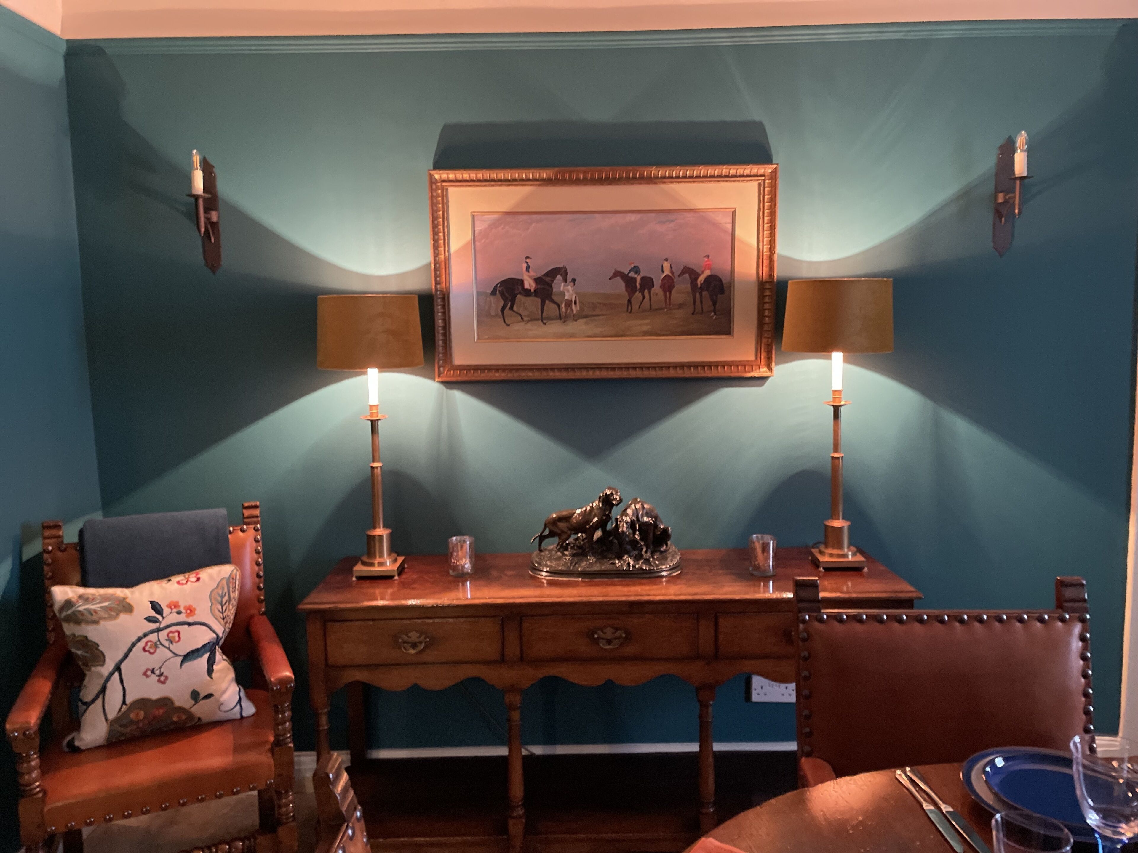 A room with a wooden desk, two lamps, a framed picture, and a chair with a floral pillow.