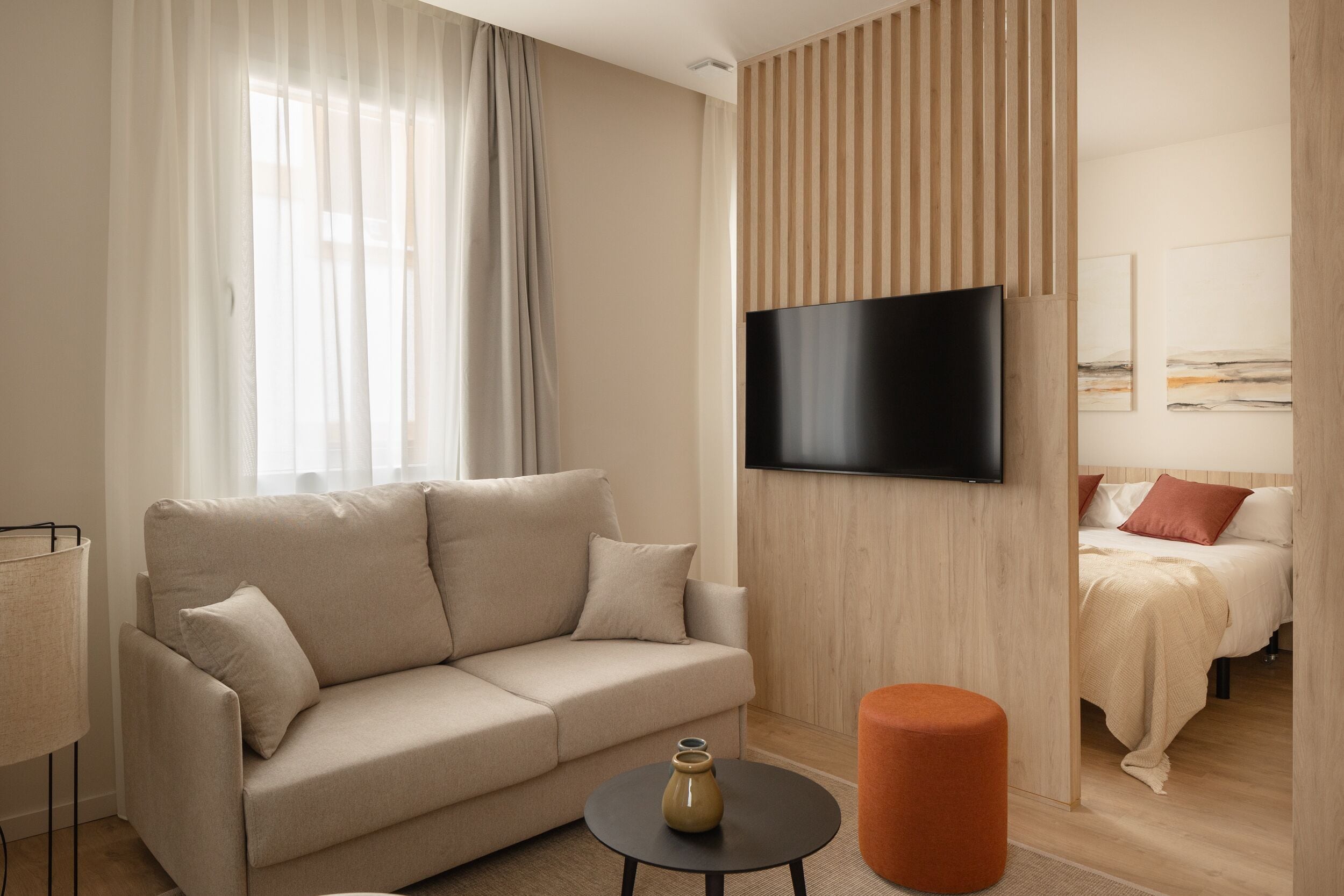 Habitació d'hotel moderna amb un sofà beix, un otomanet taronja rodó, un televisor de pantalla plana fixat a la paret i un llit amb coixins vermells.