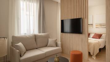 Habitació d'hotel moderna amb un sofà beix, un otomanet taronja rodó, un televisor de pantalla plana fixat a la paret i un llit amb coixins vermells.